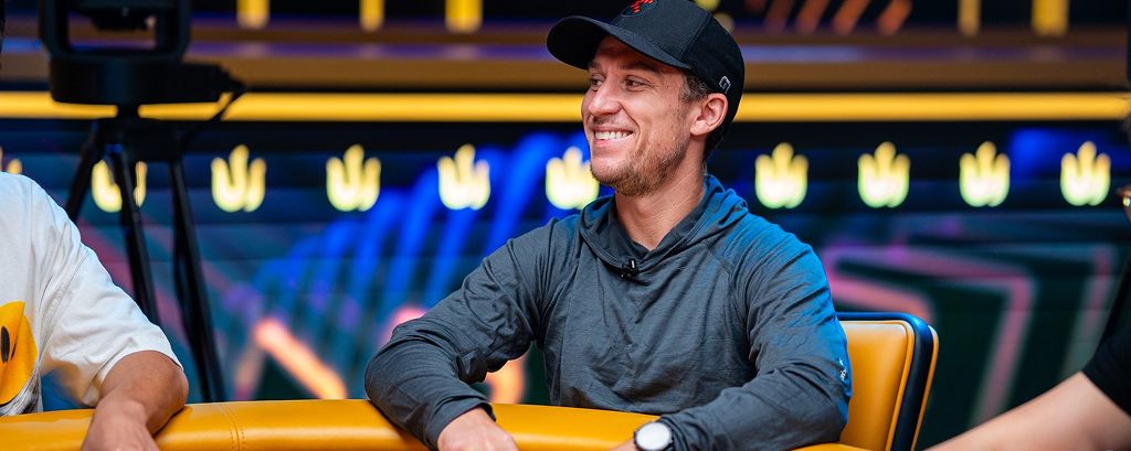 Daniel Dvoress smiling at a Triton Poker table wearing a cap and gray hoodie during a high-roller event.